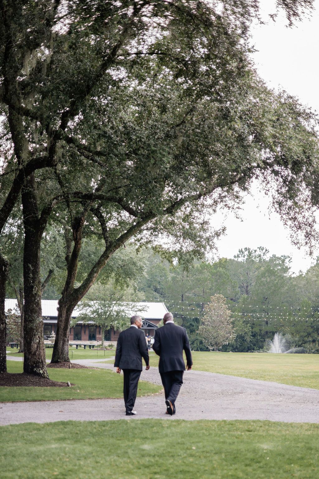 Two people in dark coats walk along a park path under trees, with a lawn and building in the background.