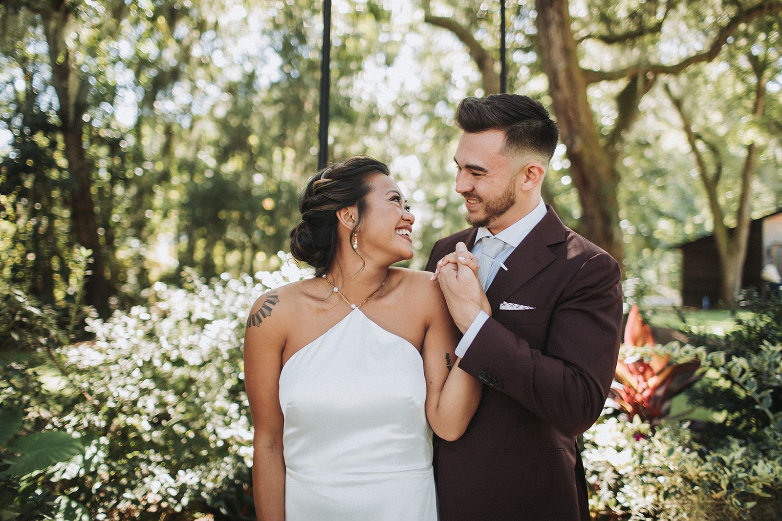 Bride and groom smiling in a garden, holding hands amid greenery and white flowers