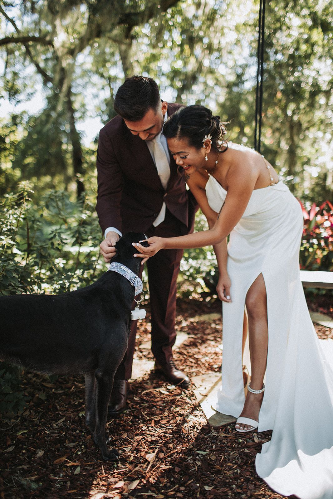 Bride in white dress and groom in suit petting a black dog outdoors under trees