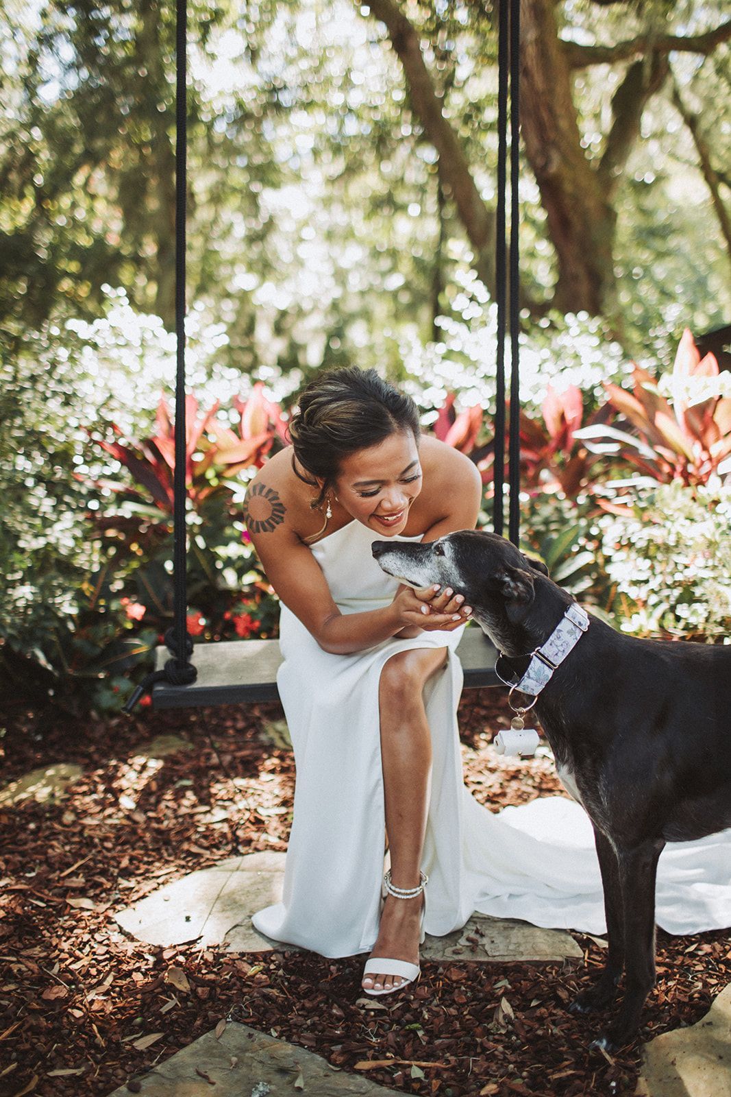 Bride in a white dress kneels outdoors, smiling at a black dog in a garden setting.