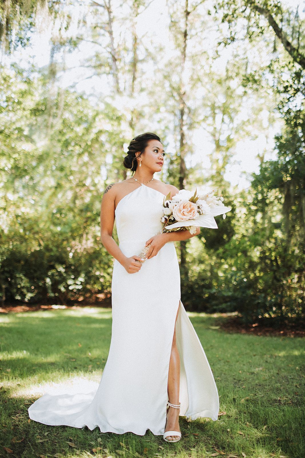 Bride in a white gown holding a bouquet outdoors beneath sunlit trees