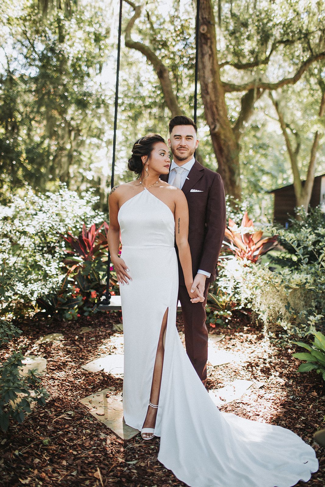 Couple in formal attire standing together in a sunlit garden, bride in white gown with long train