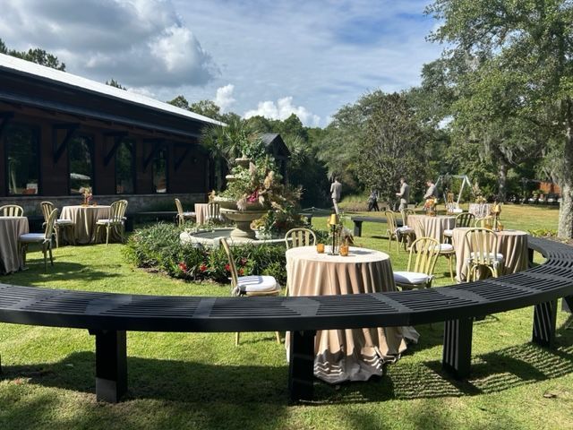Outdoor garden reception with round tables, beige linens, and a curved black bench under a partly cloudy sky