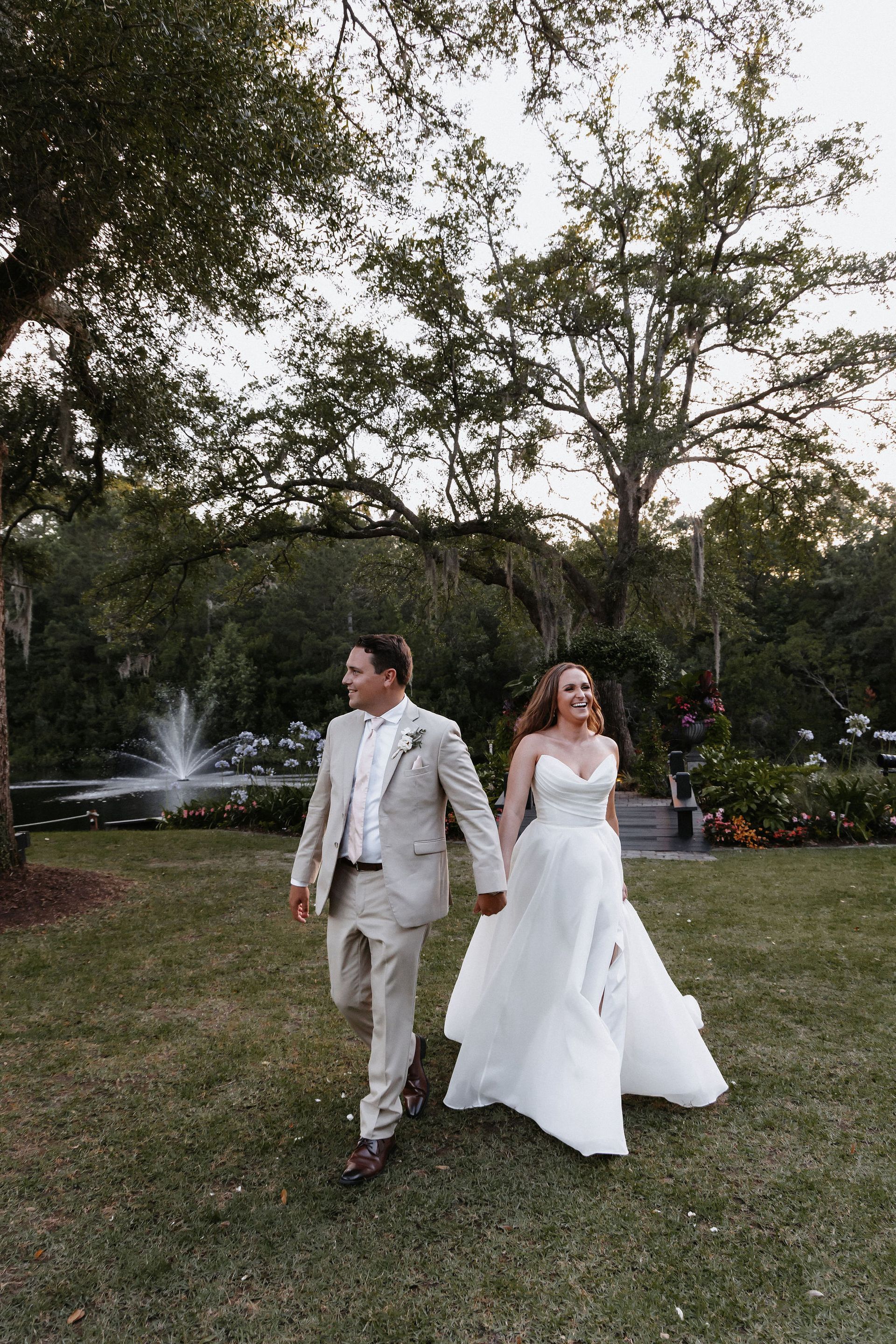 Couple in wedding attire walking on grass in a garden with a fountain behind them.