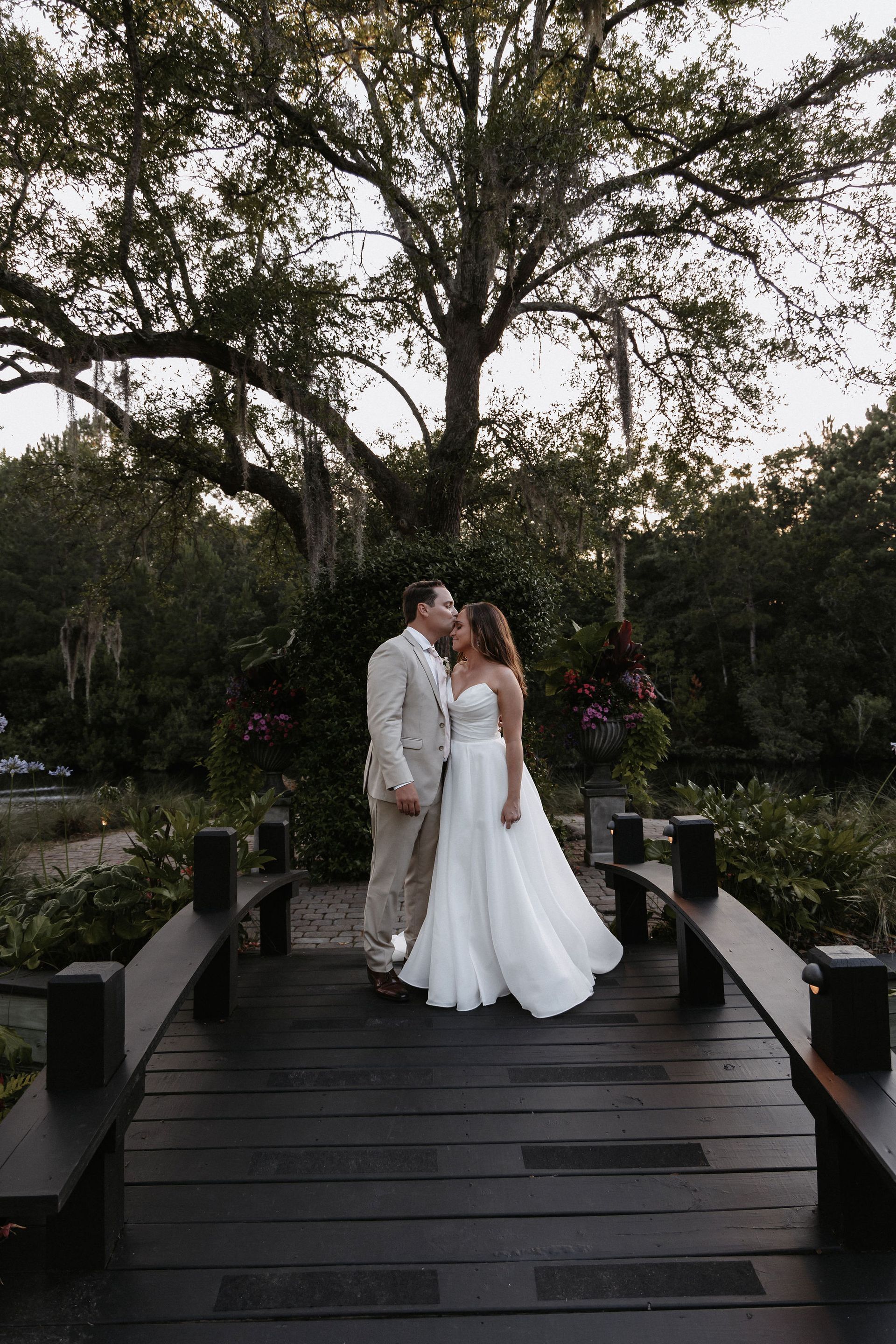 Couple in white wedding attire standing on a wooden bridge in a garden at dusk.