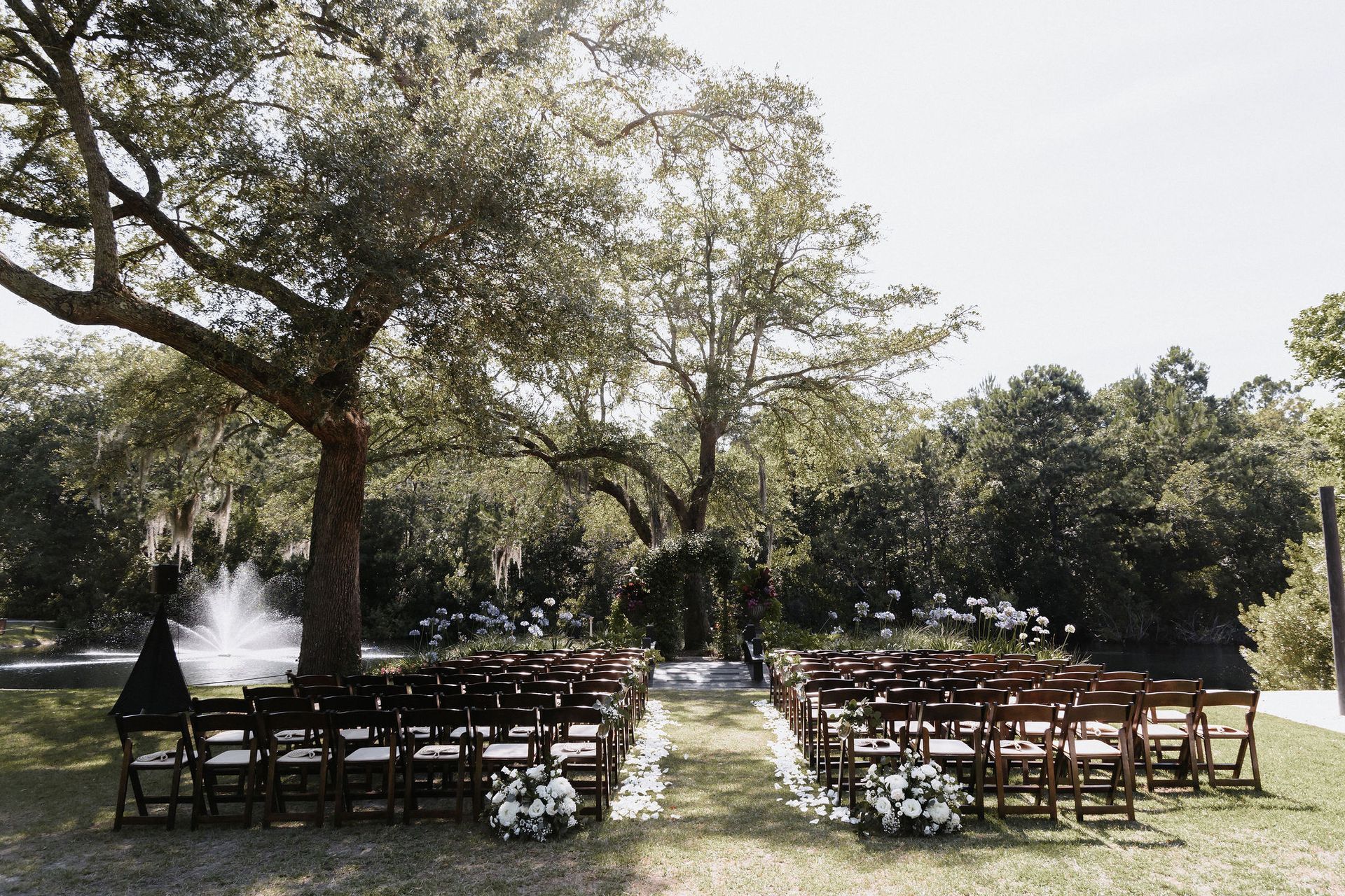 Outdoor wedding setup with rows of wooden chairs under trees on a sunny lawn