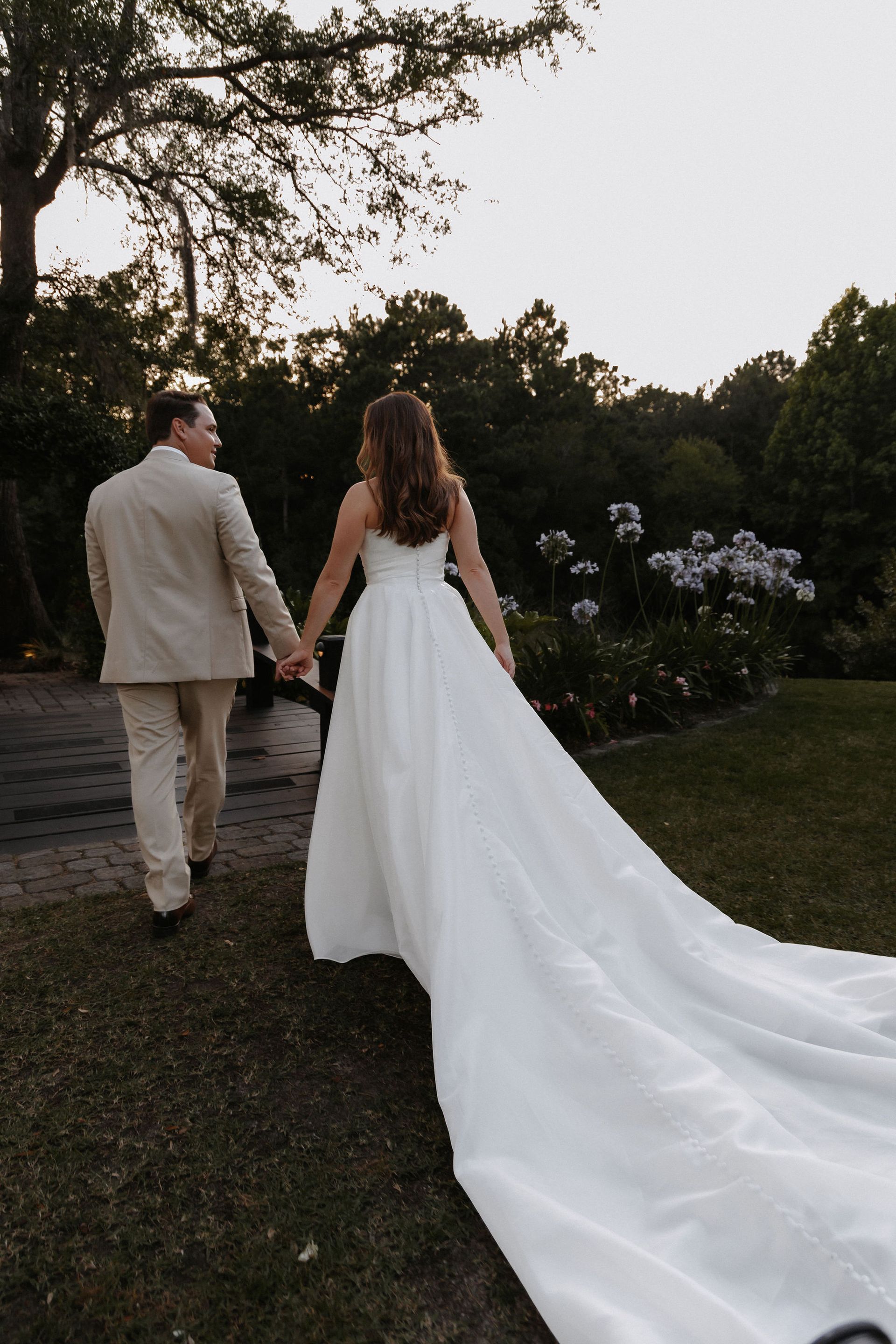 Newlyweds walking hand in hand down a garden path, bride in a long white dress and groom in a beige suit