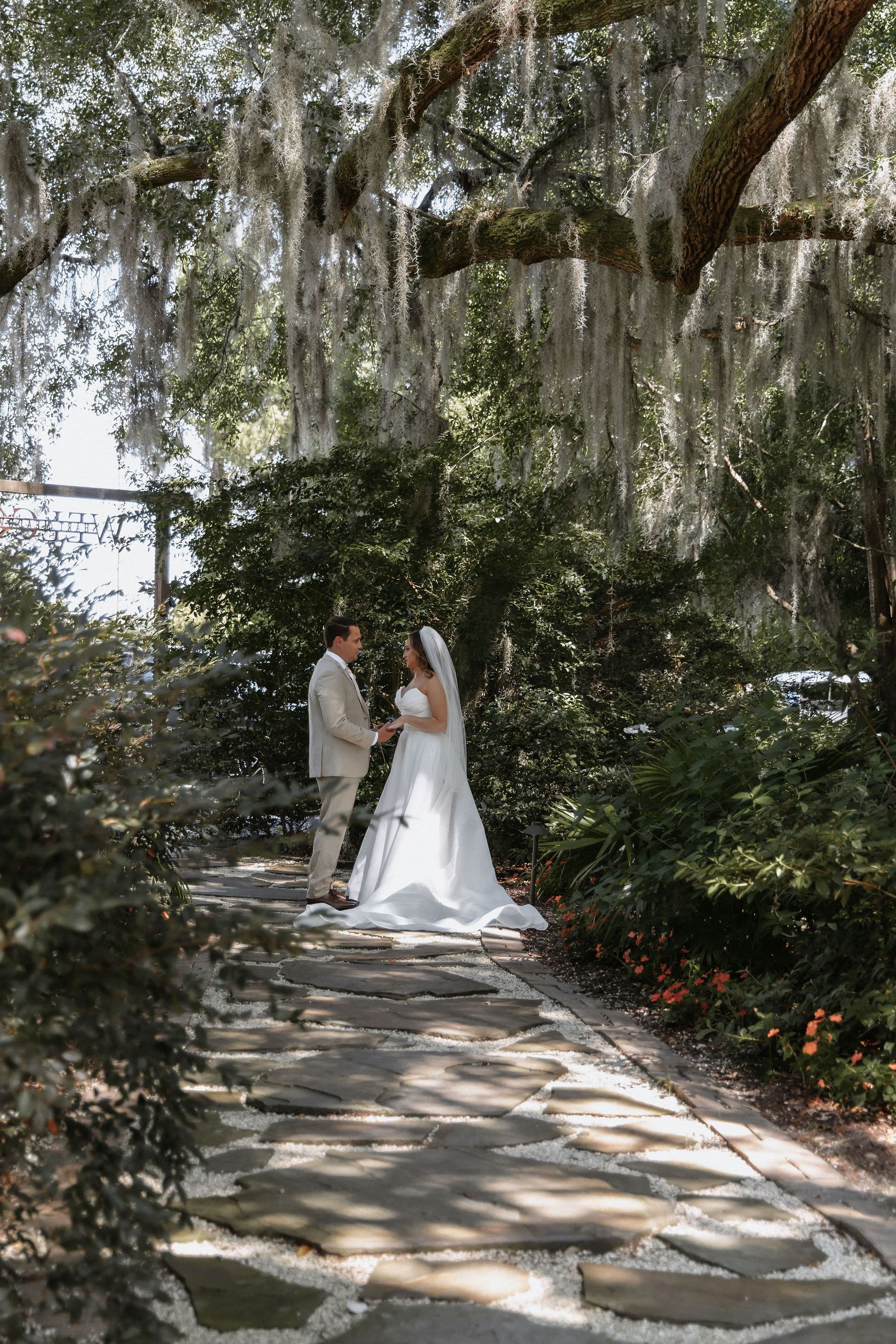 Bride and groom standing on a stone path beneath hanging moss in a garden setting