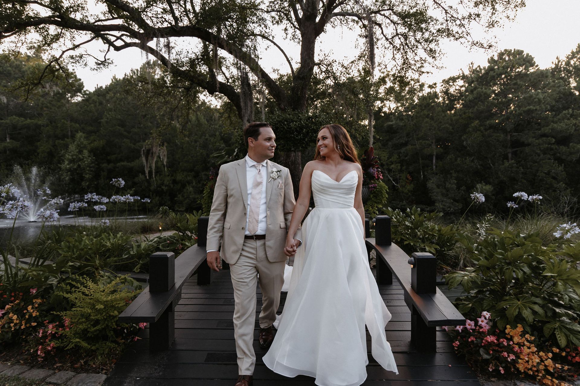 Bride and groom walking hand in hand across a garden bridge at dusk, surrounded by flowers and trees