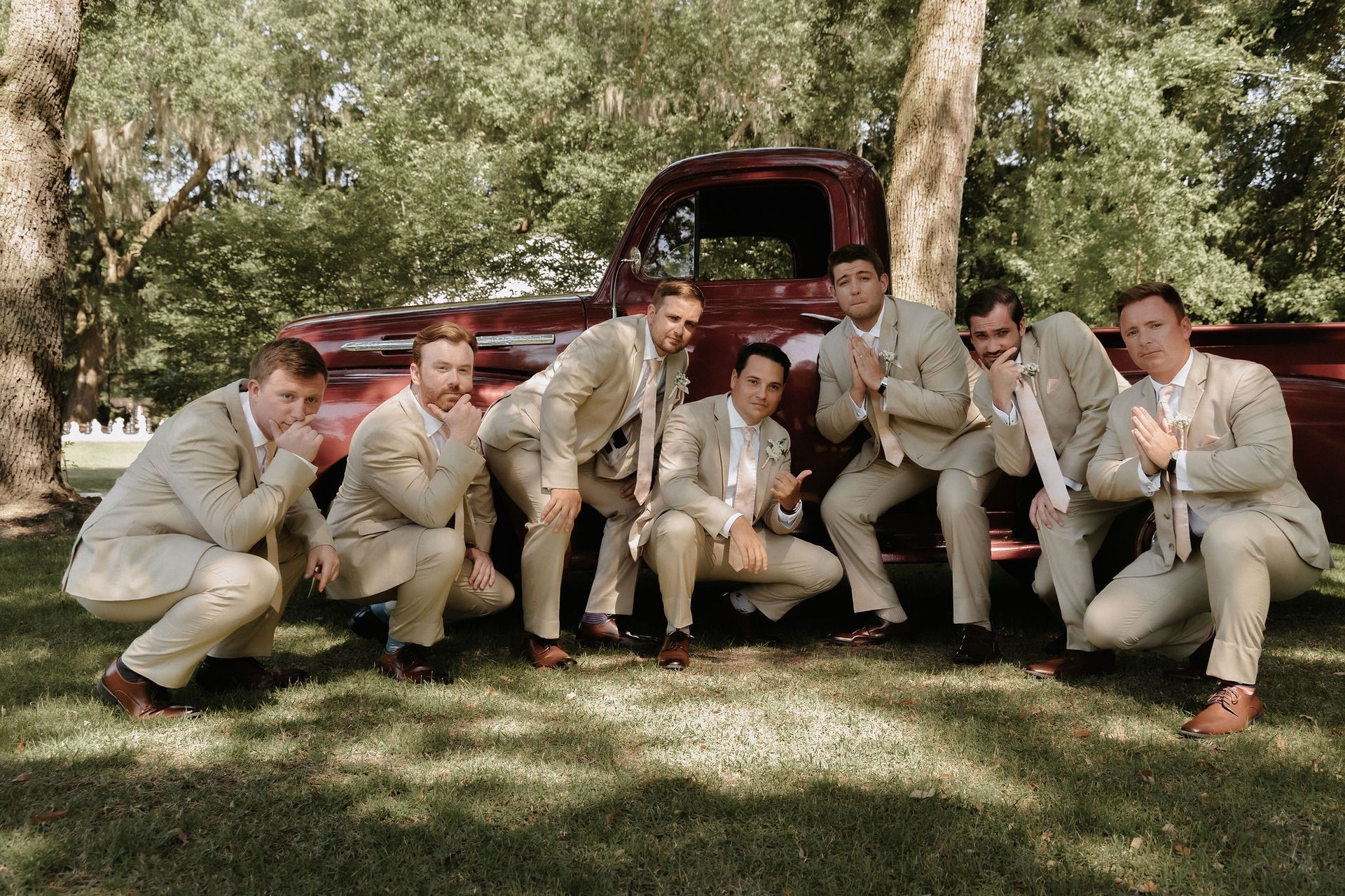 Group of people in beige suits posing outdoors beside a red vintage truck under trees.