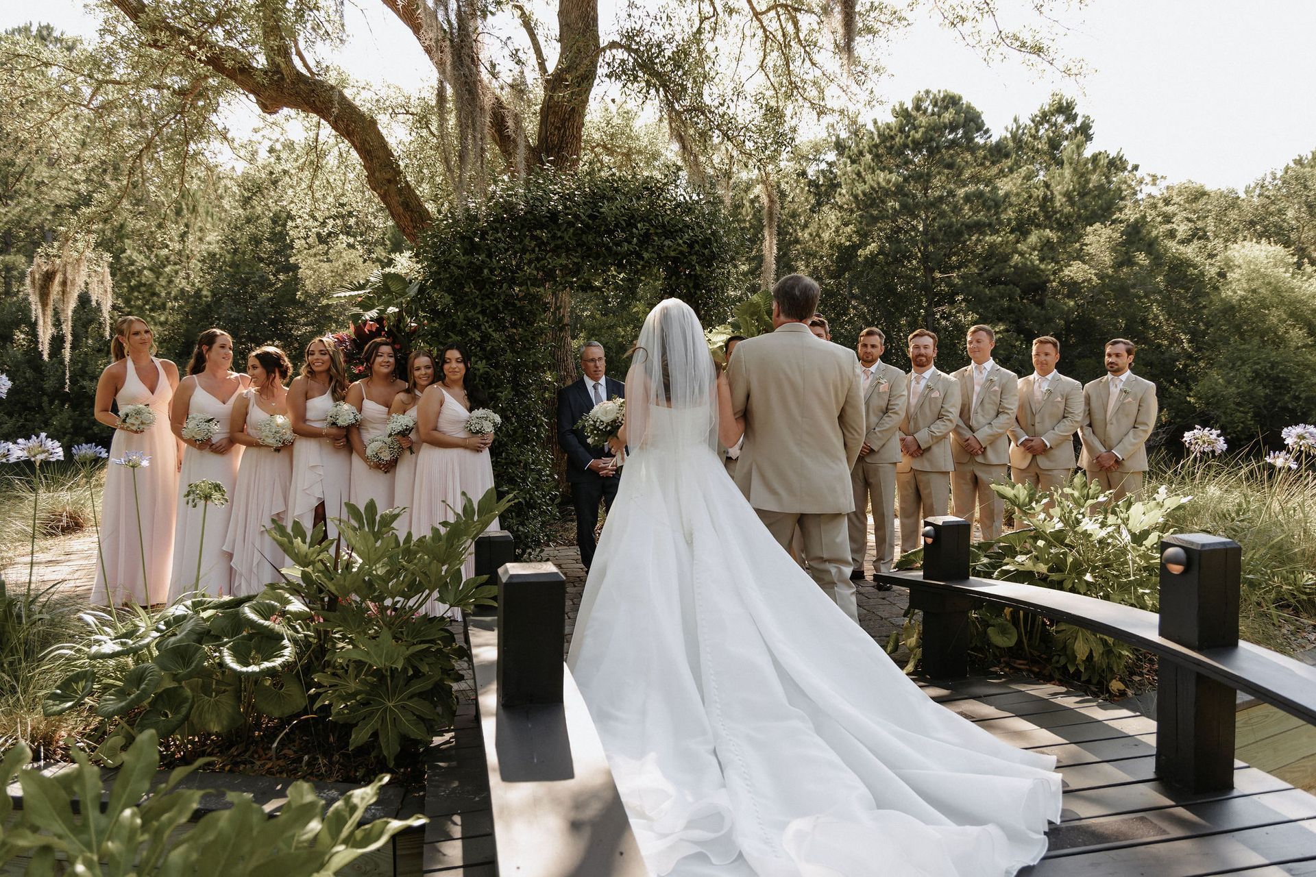 Wedding party on a garden bridge, bride in white gown facing guests and groom in tan suit