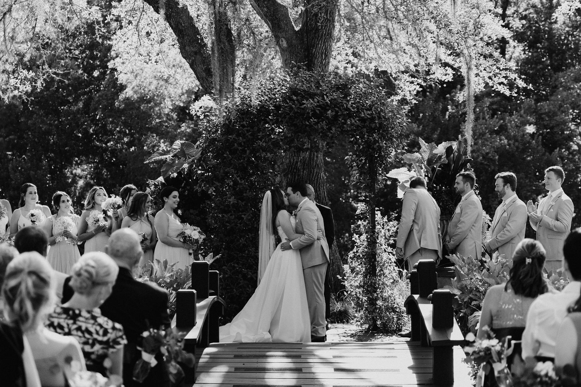 Bride and groom exchange vows at an outdoor wedding ceremony under trees, guests seated nearby.