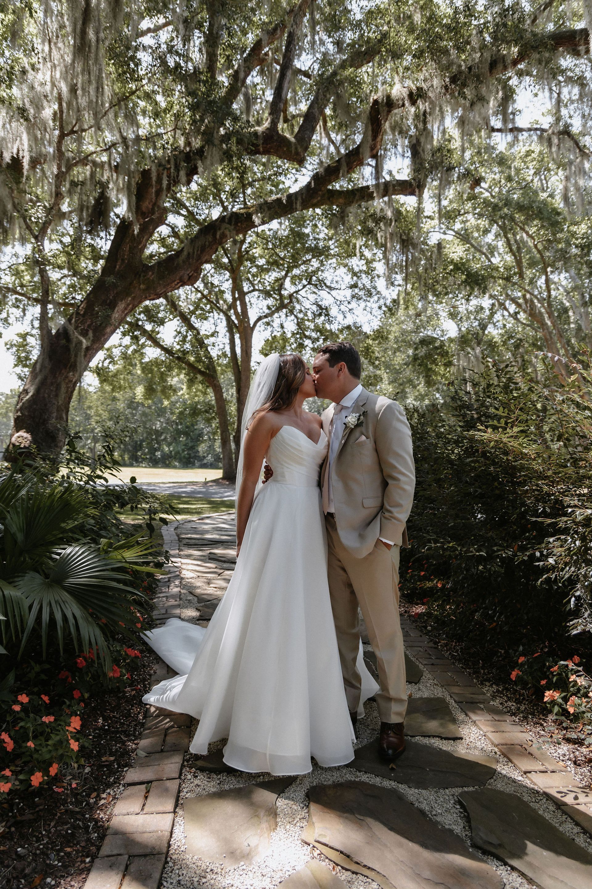 Bride and groom kissing under a large tree in a garden path, bride in white gown and groom in tan suit