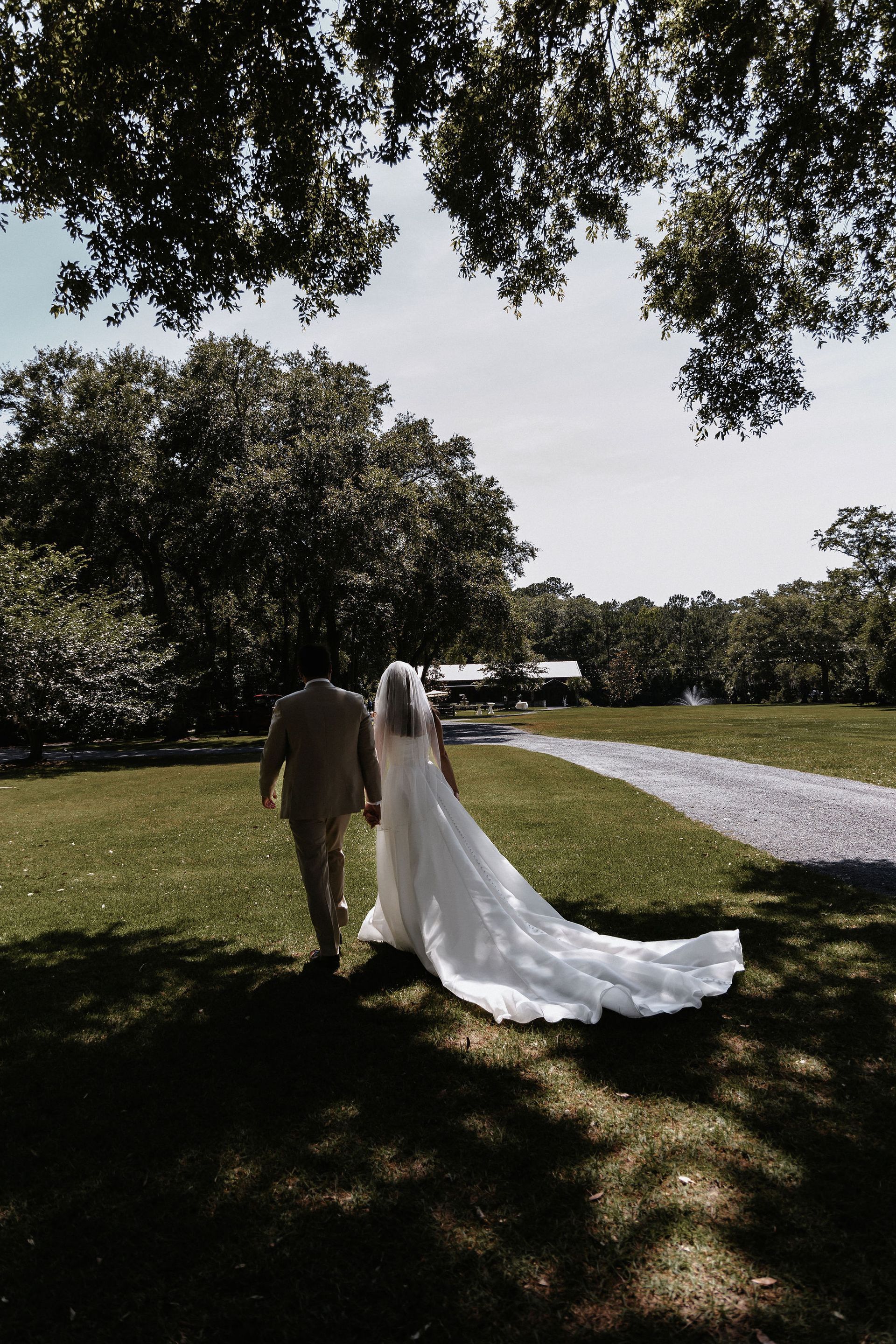 Bride and companion walking on a grassy path, with a long white veil and train in a sunny park.