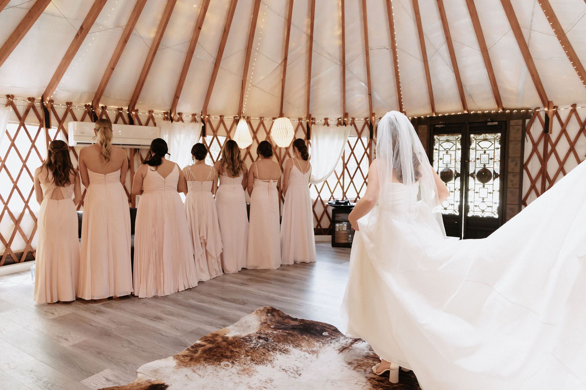 Bridesmaids in blush dresses stand in a yurt as a bride in a long veil faces them from the right