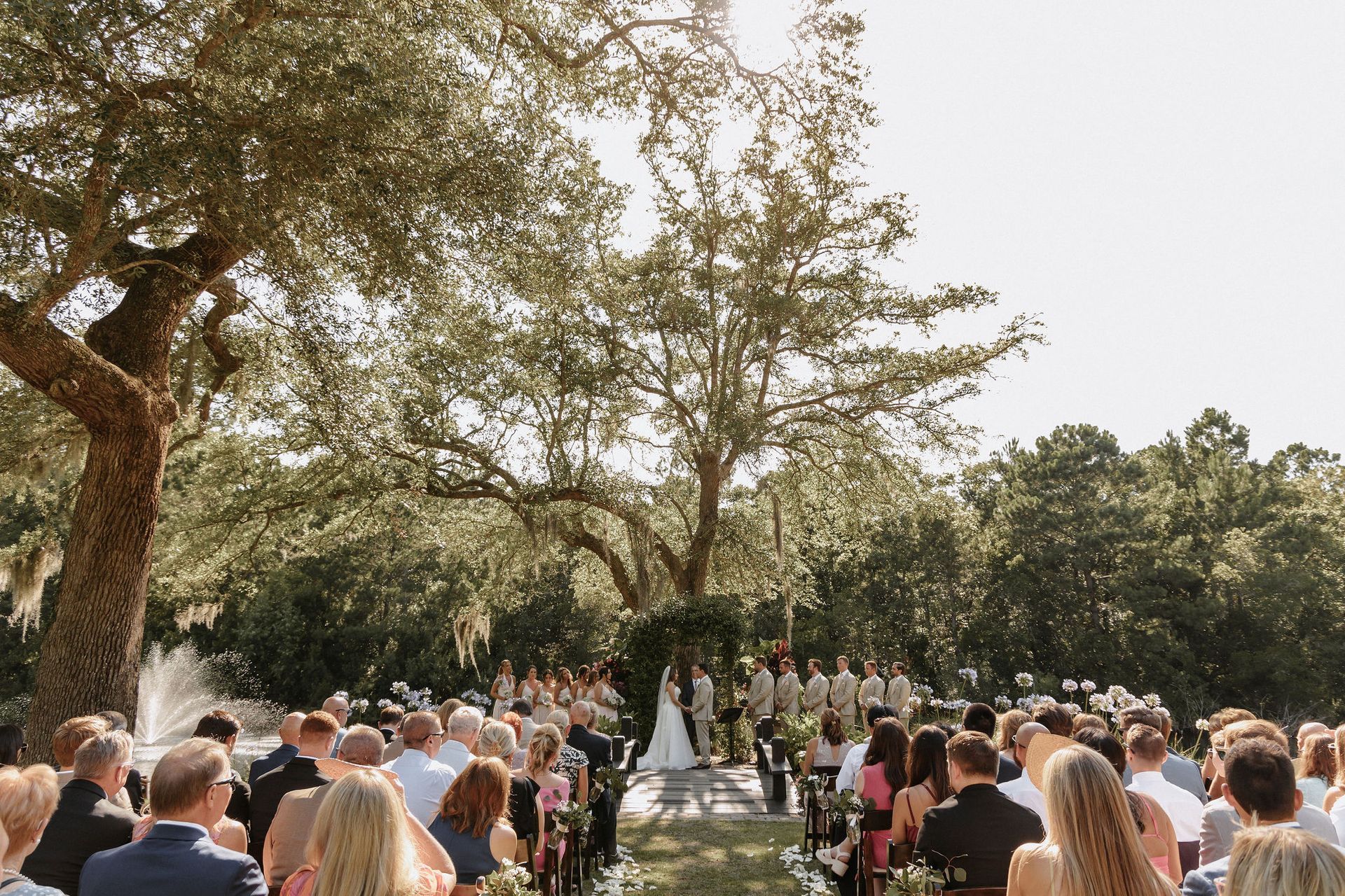 Outdoor wedding ceremony under trees with guests seated along a white aisle at sunset