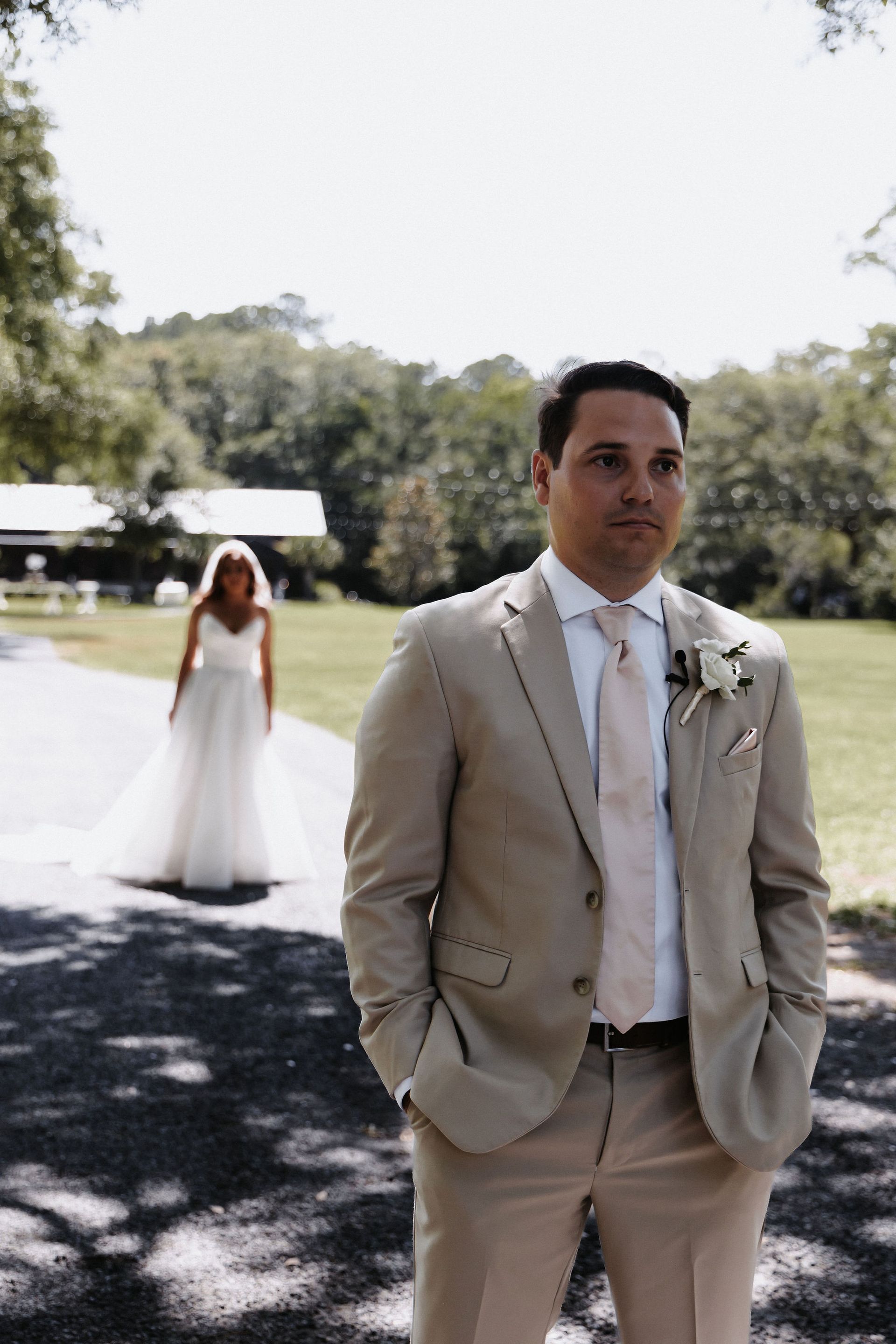 Man in beige suit stands on a path; bride in white dress walks behind him in a sunny park.
