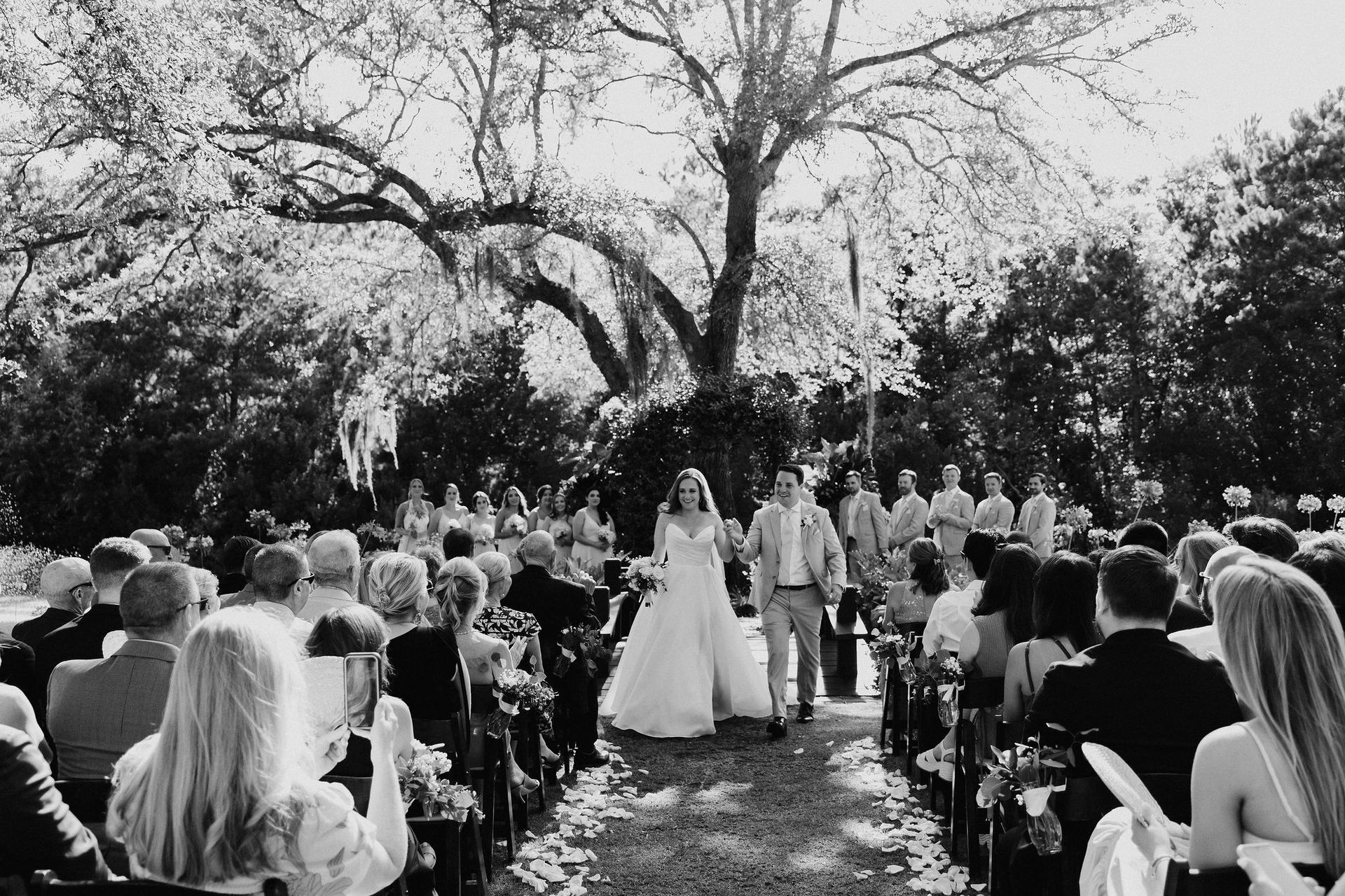 Outdoor wedding ceremony under a large tree, with bride and groom walking down an aisle between seated guests