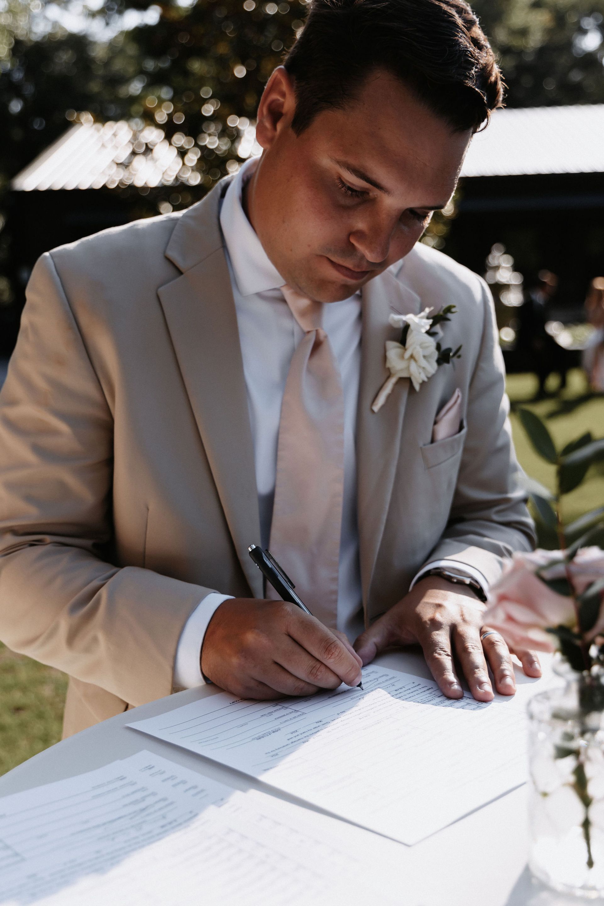 Man in a light gray suit signing a document at an outdoor wedding ceremony
