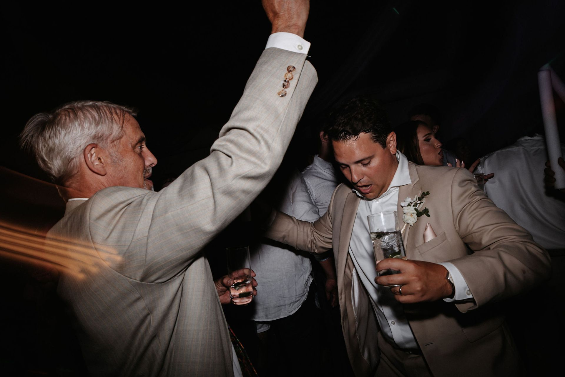 People dancing at a dimly lit party, one raising an arm while holding drinks.