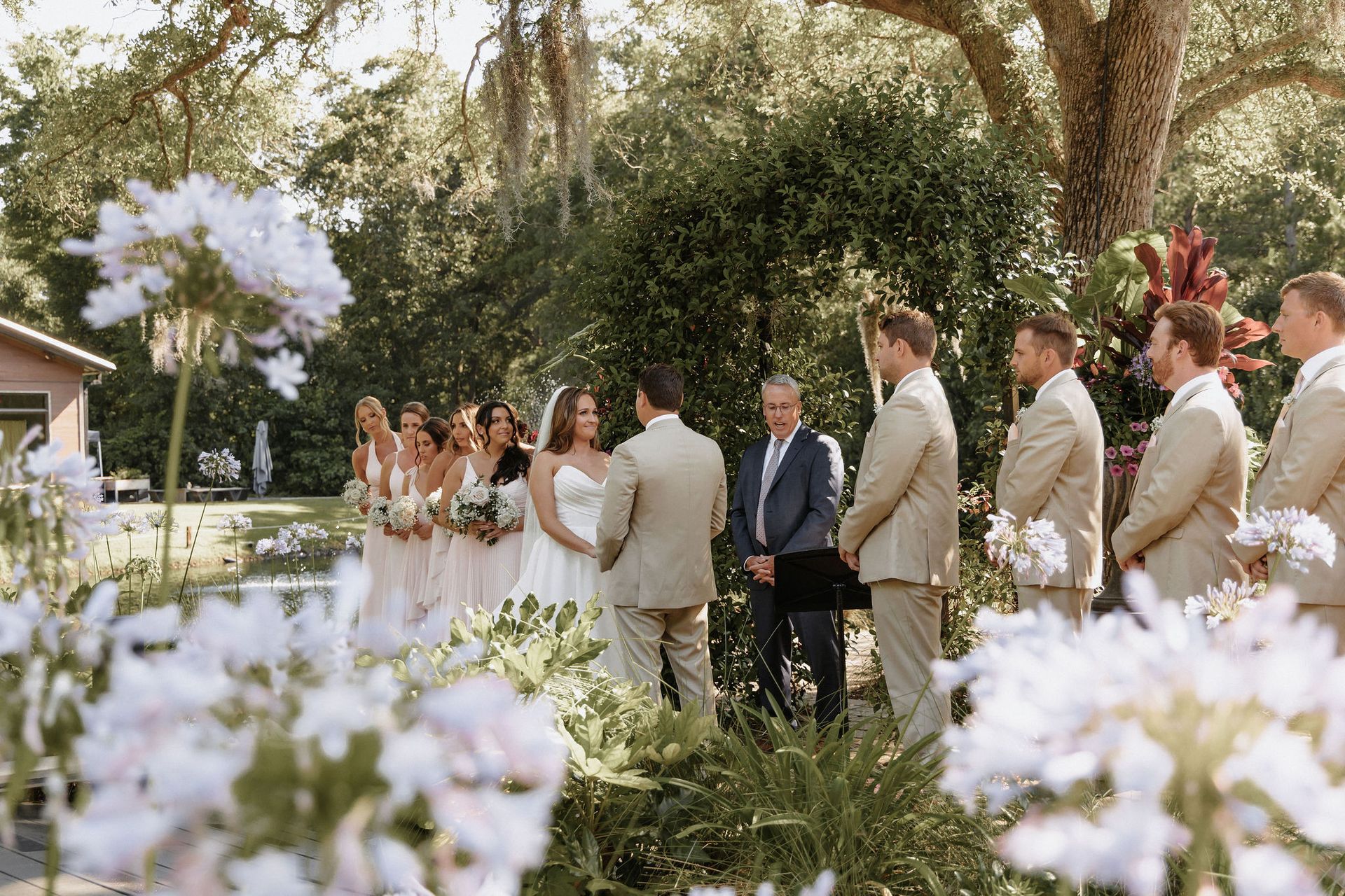 Wedding party standing in a garden ceremony, framed by white flowers and greenery.