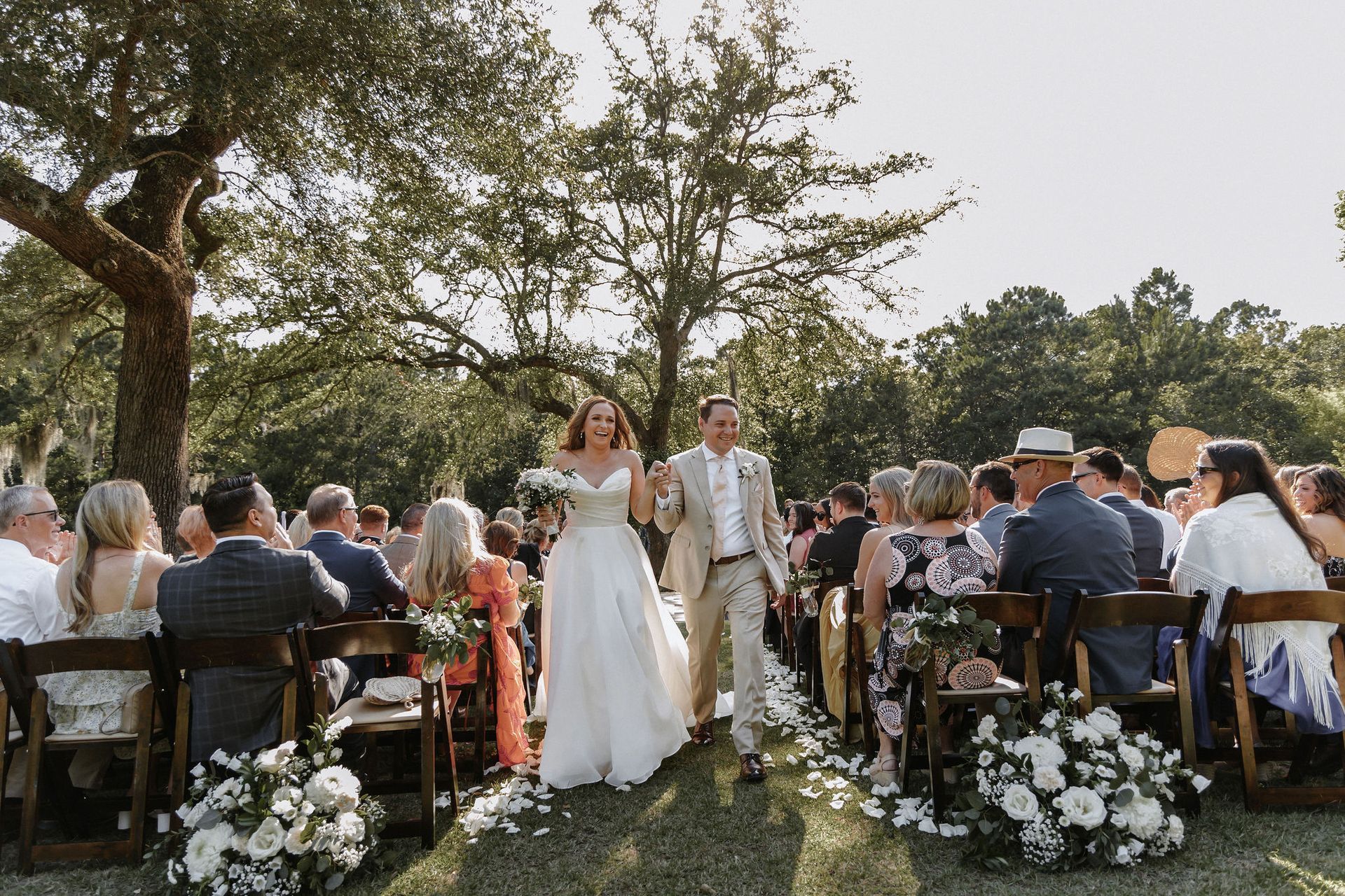 Wedding couple walking down an outdoor aisle between seated guests under trees