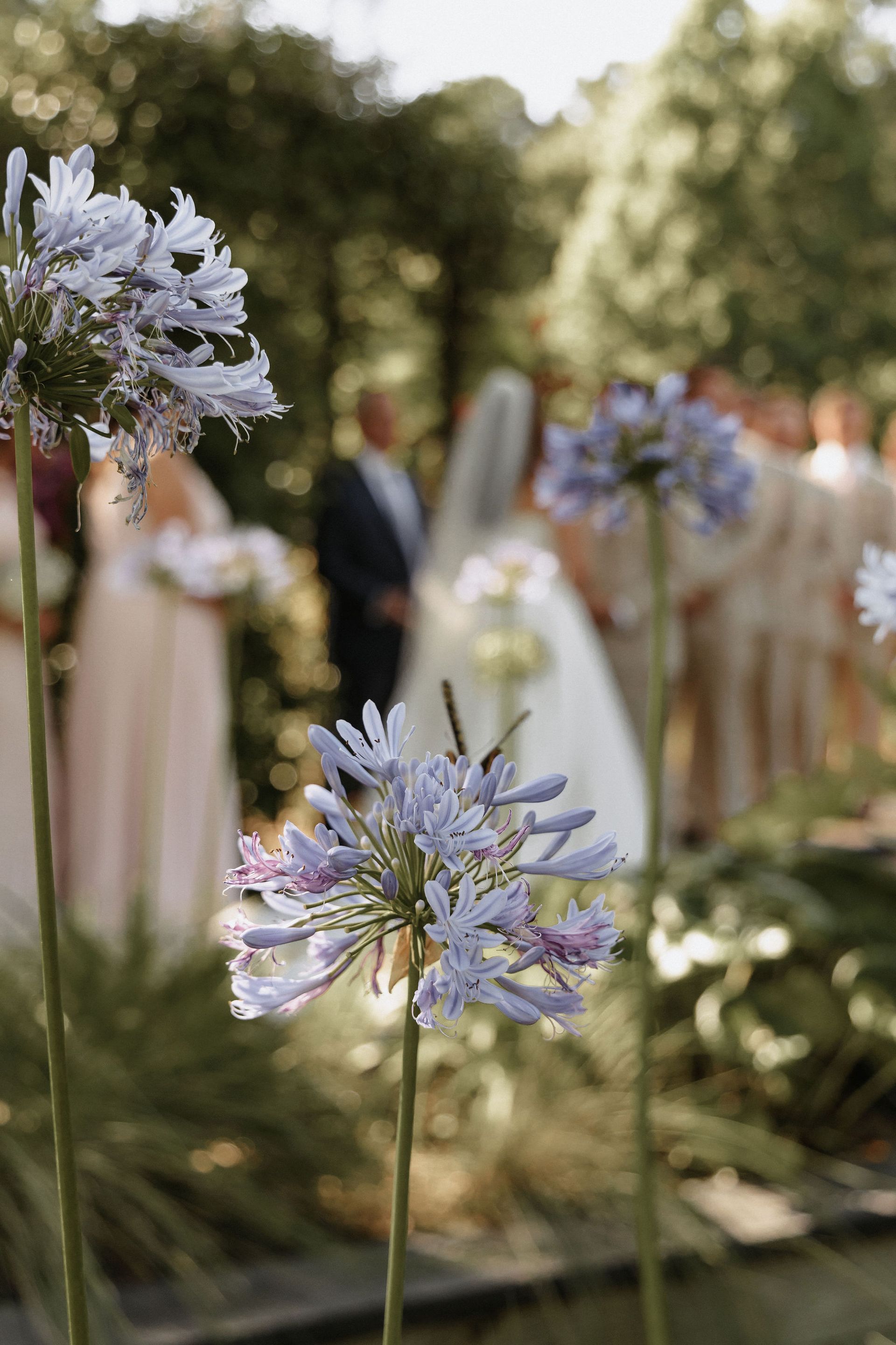 Purple flowers in focus at an outdoor wedding, with blurred guests and white floral arrangements in the background