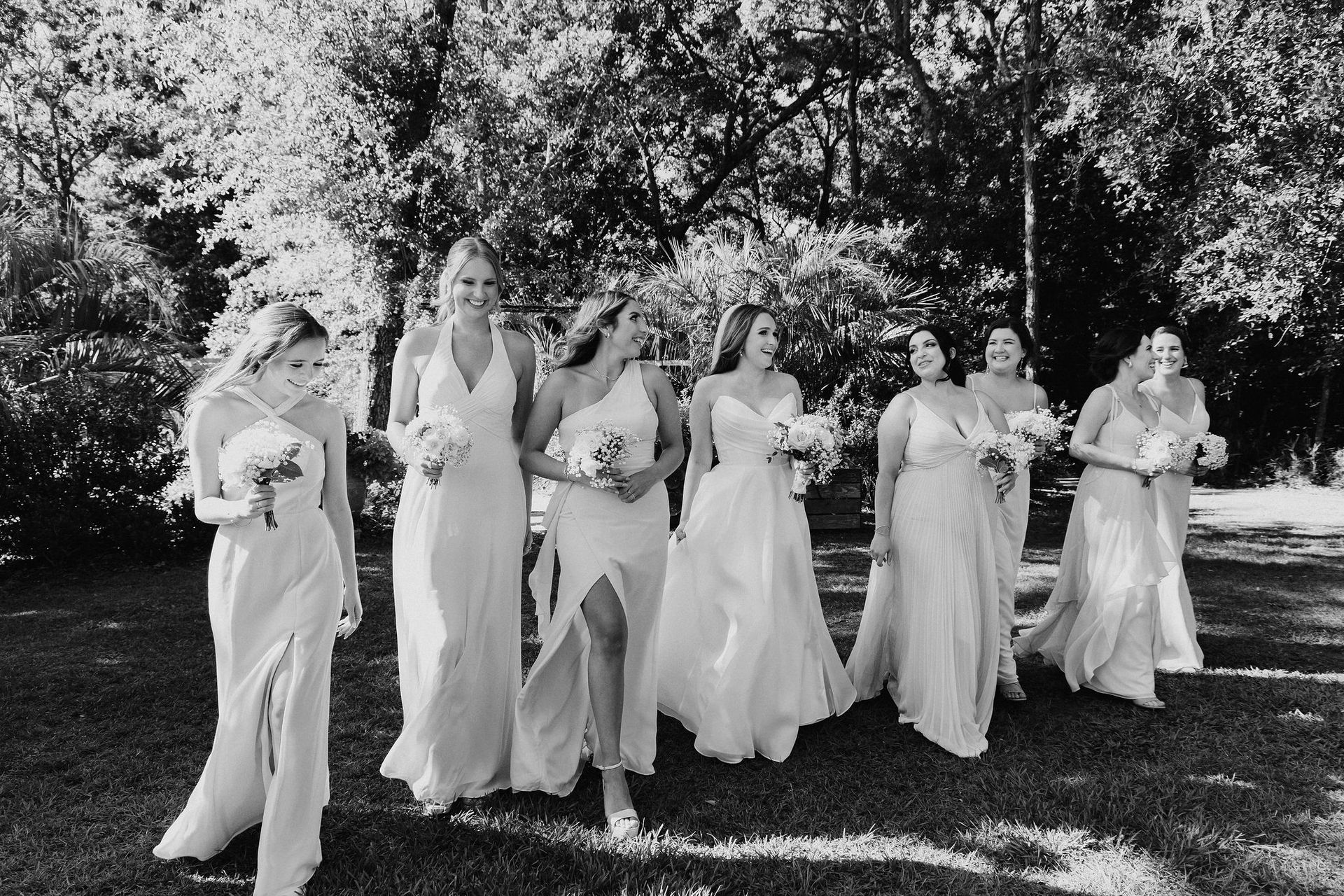 Six women in white dresses walk together outdoors under trees, holding bouquets.