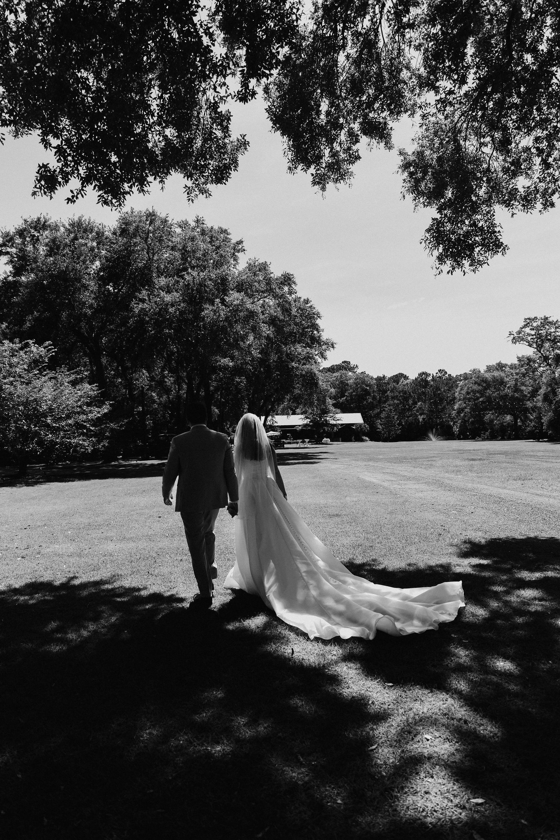 Two people walking under trees on a sunny lawn, one in a long veil or dress trailing behind.