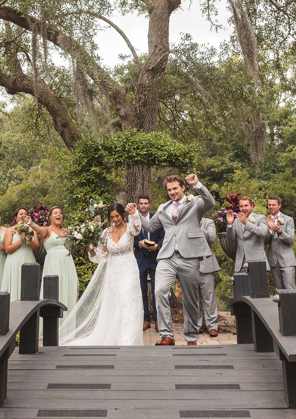 Wedding party gathered on a wooden bridge outdoors, with a bride and groom at center, cheering and clapping.