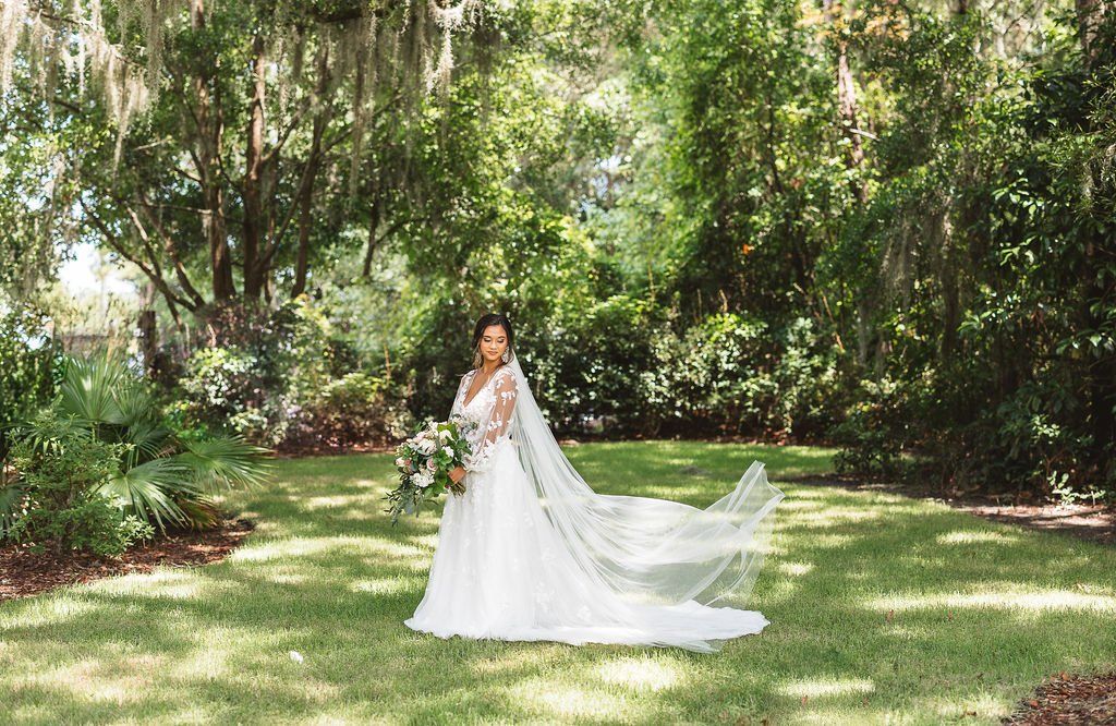 Bride in a white gown holding a bouquet in a sunlit garden with flowing veil train