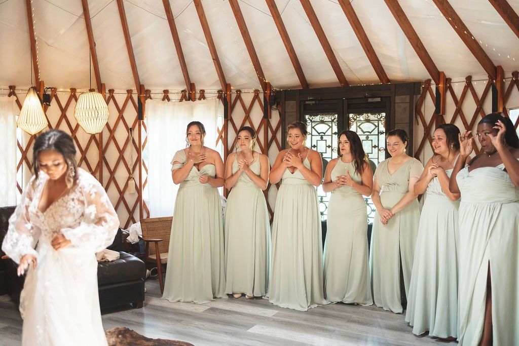 Bride in white dress with bridesmaids in pale green dresses inside a rustic yurt, clapping and smiling