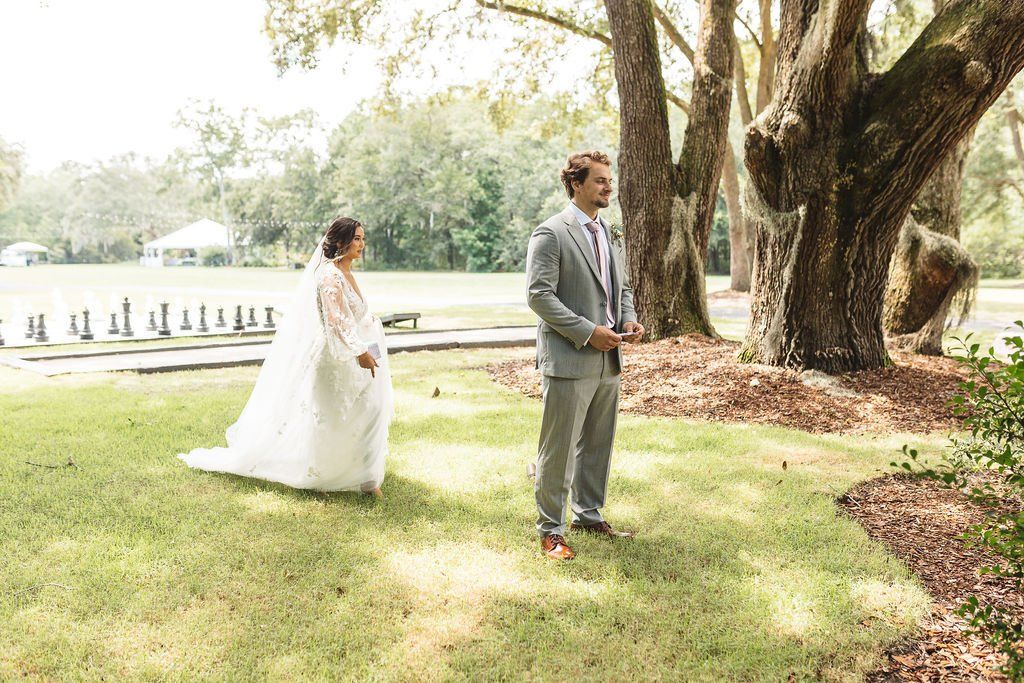 Bride in white dress and groom in gray suit walking on a sunny lawn near large trees and water