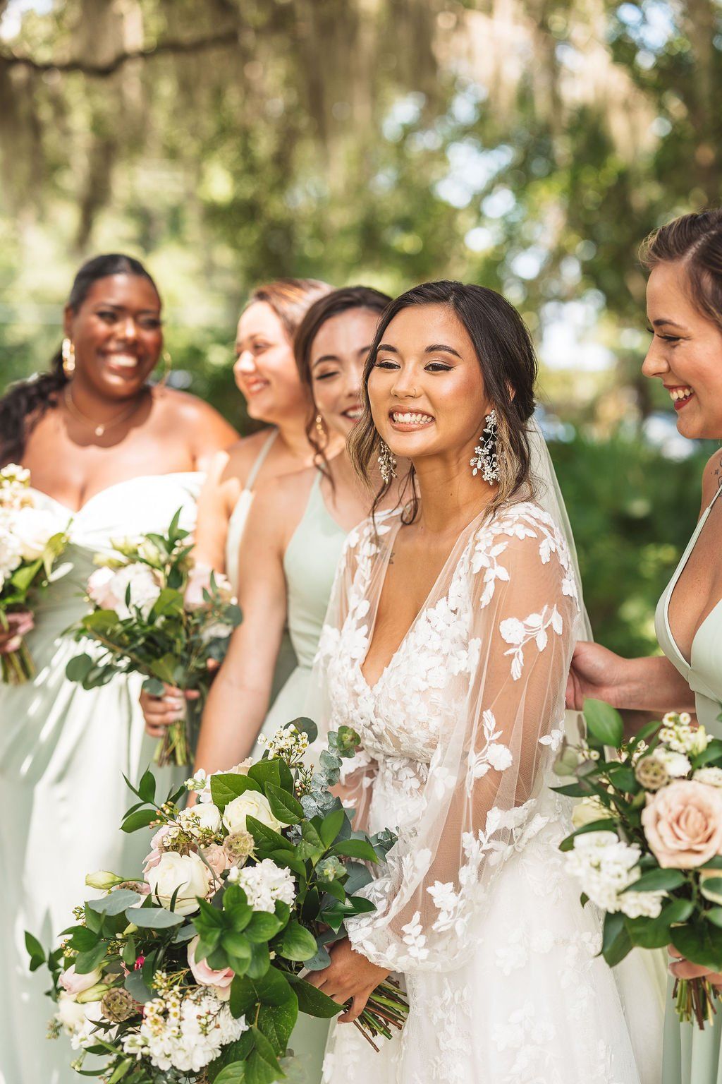 Bride in a white lace dress with bridesmaids holding bouquets outdoors among greenery