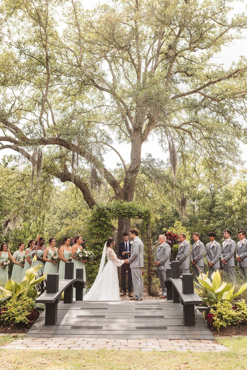 Wedding ceremony outdoors on stone steps beneath a large tree, with guests and wedding party lined on both sides.