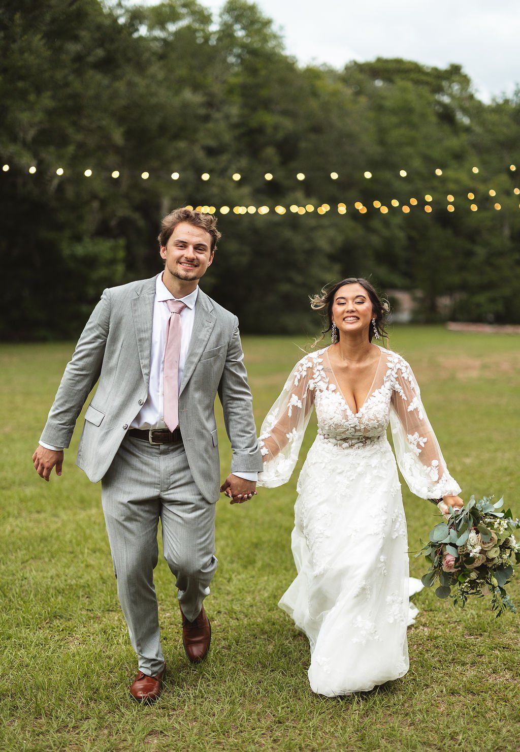 Bride and groom walking hand in hand on grass under string lights, smiling in a garden wedding ceremony