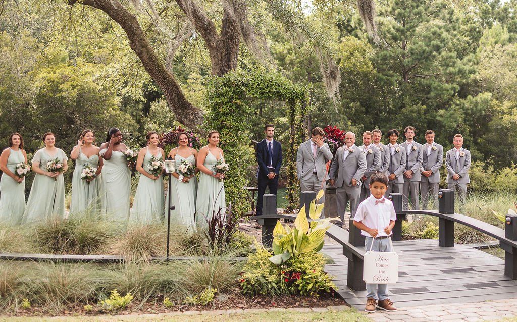 Group of people in formal attire standing in a garden beside a walkway and bridge