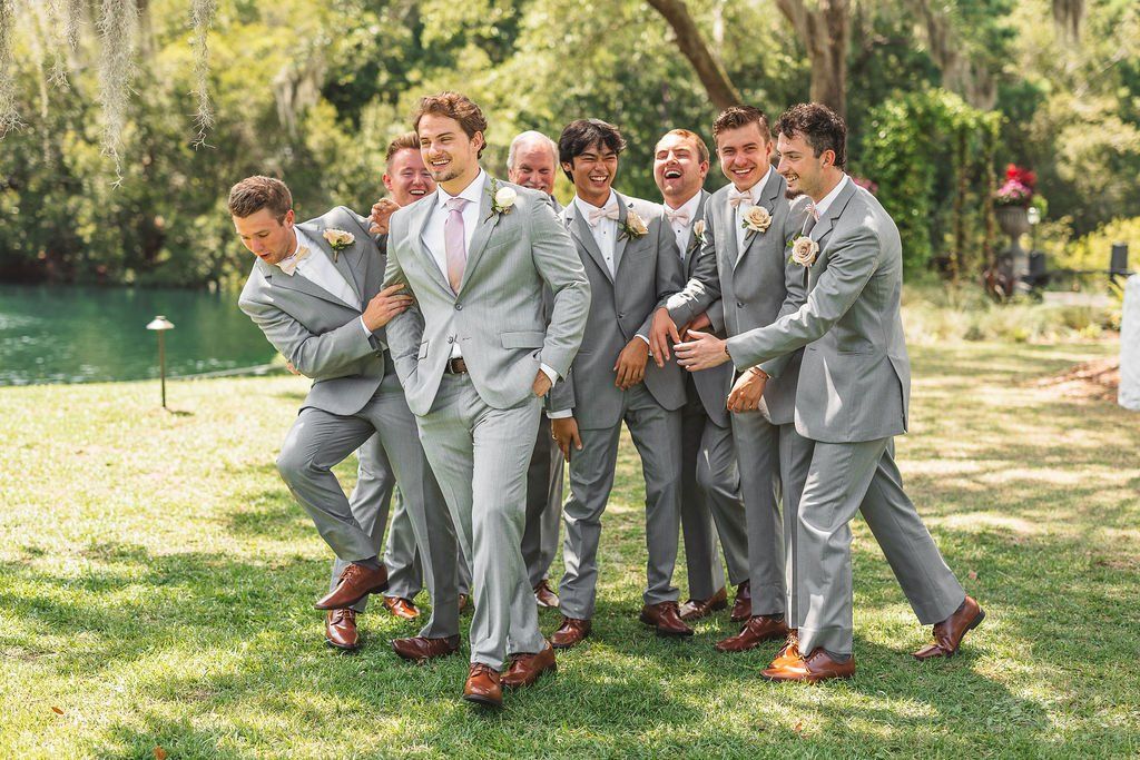 Group of six men in gray suits laughing and walking together on a sunny lawn near a pond