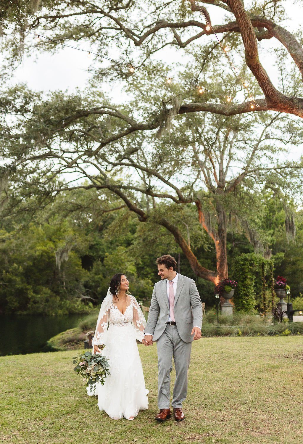 Couple in wedding attire walking hand in hand through a grassy park beside a lake