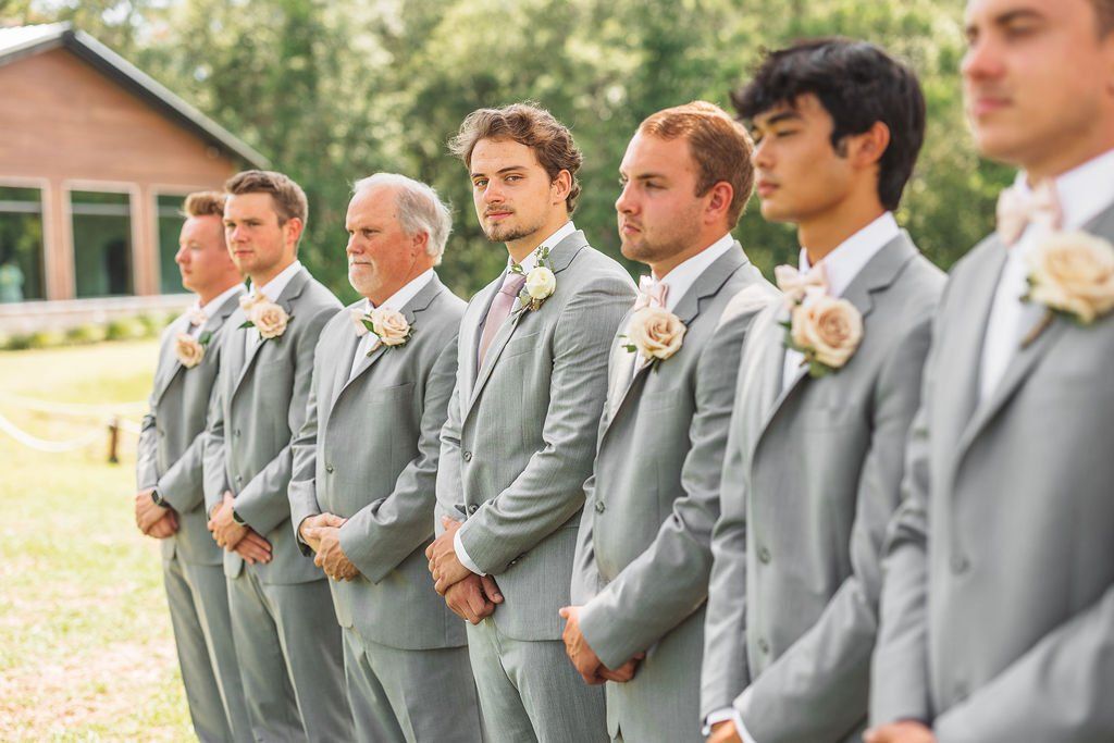 Groomsmen in light gray suits stand in a row outdoors at a wedding ceremony.