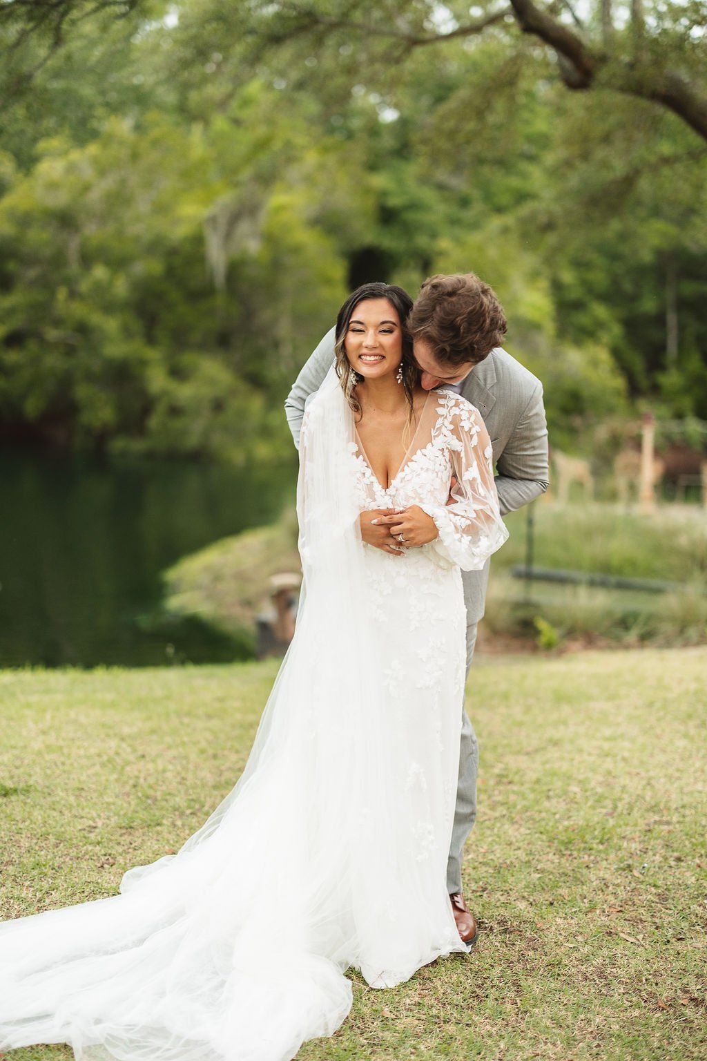 Bride in a white lace gown and veil smiles outdoors as a partner stands behind her on a grassy lawn.