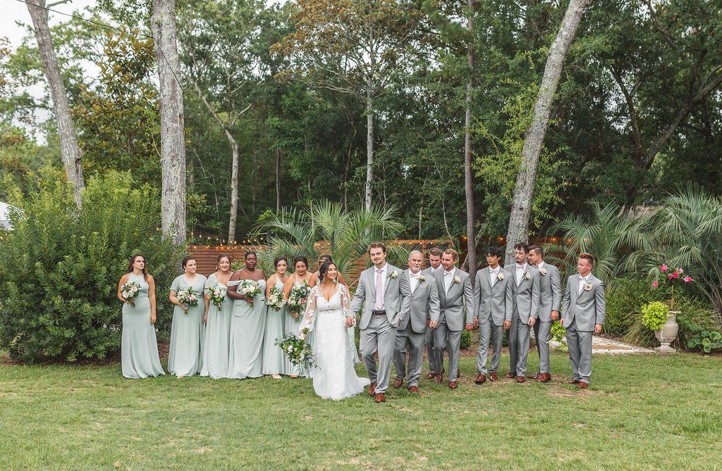 Wedding party posing outdoors in gray suits and sage dresses with bride in white, surrounded by greenery