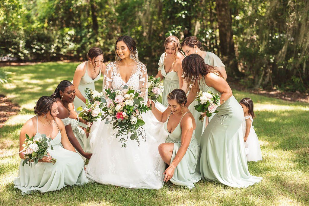 Bridesmaids in sage green dresses gather around a bride in a white gown holding a bouquet outdoors on grass.