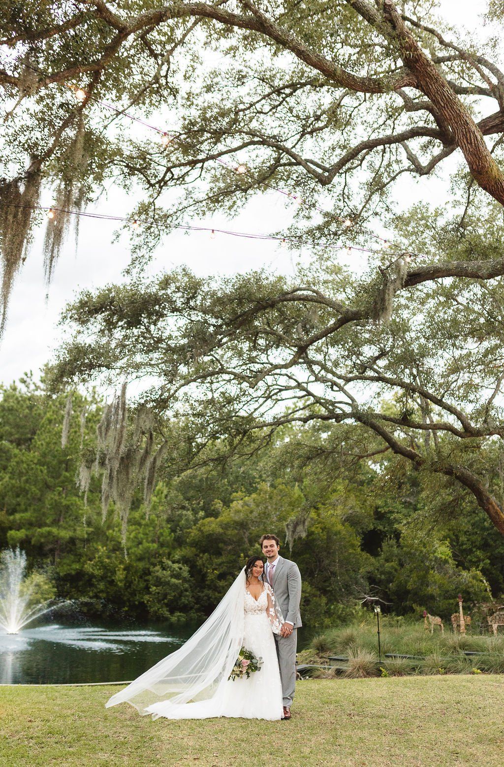 Bride and groom standing in a garden under a large tree, with a fountain in the background