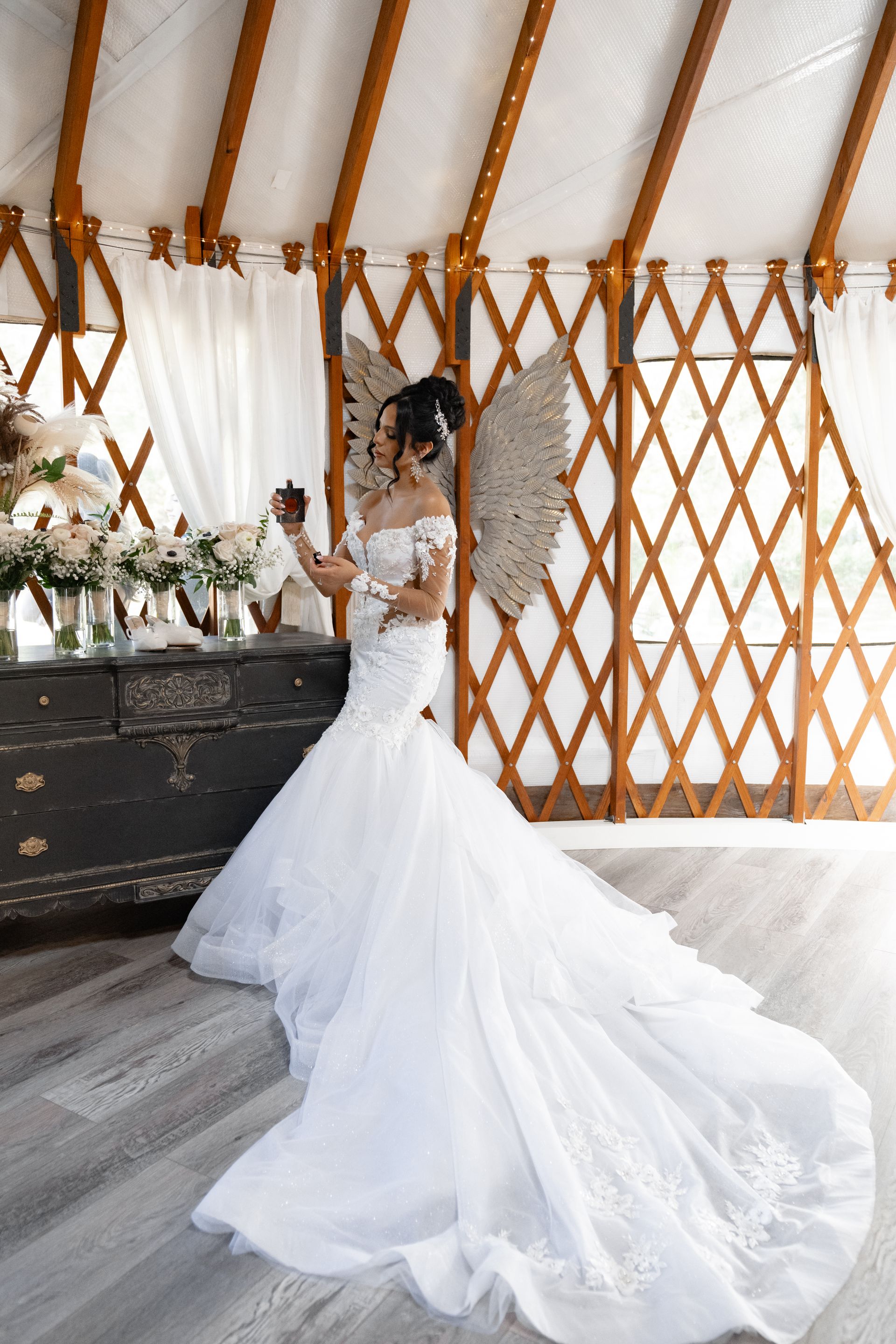 Bride in a white wedding gown standing beside a dark dresser in a decorated tent with wooden lattice walls.