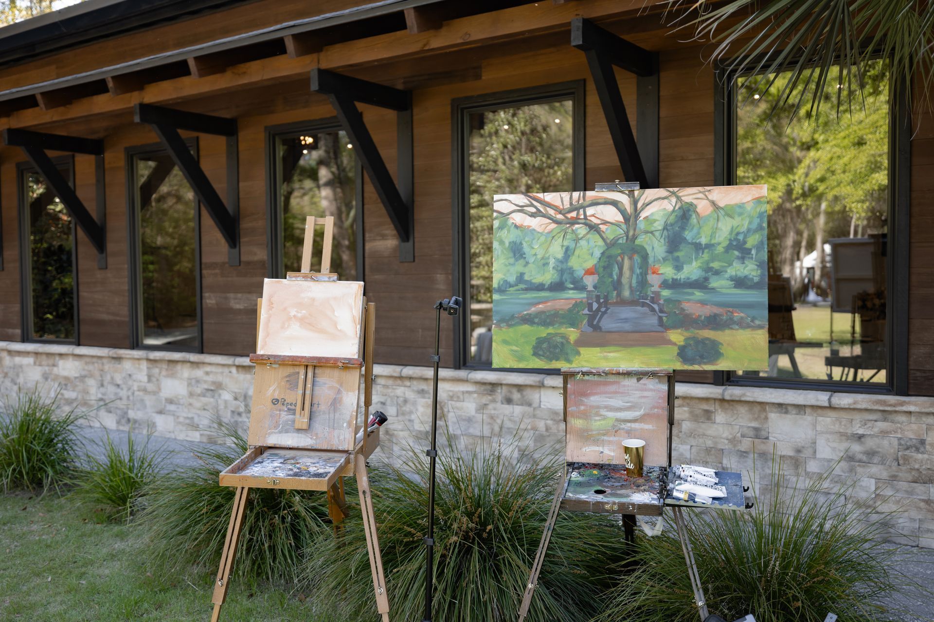 Two easels with paintings set outside a rustic building, with greenery and windows behind them.