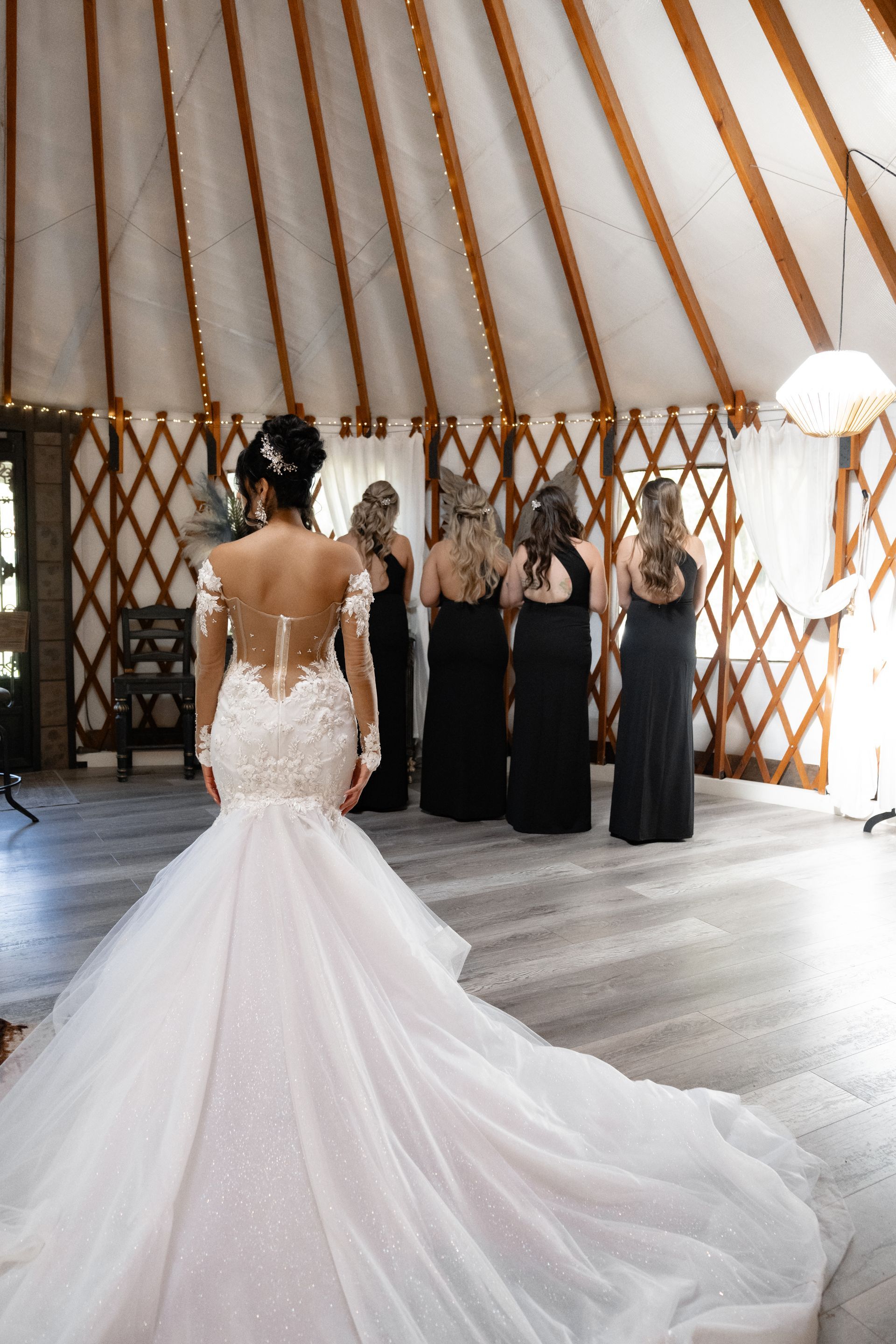 Bride in a white gown facing bridesmaids in black dresses inside a rustic tented venue