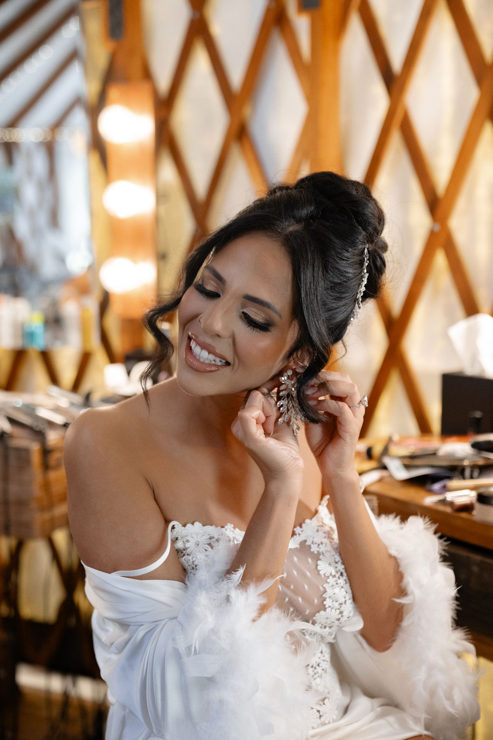 Smiling bride in a white off-shoulder dress adjusting her earring in a warmly lit room