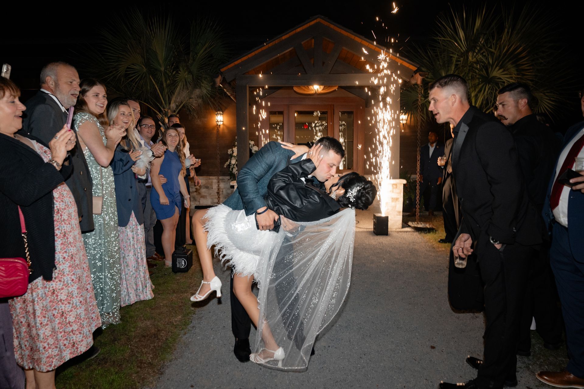 Bride and groom kiss at nighttime wedding send-off as guests line the walkway and fireworks sparkle behind them