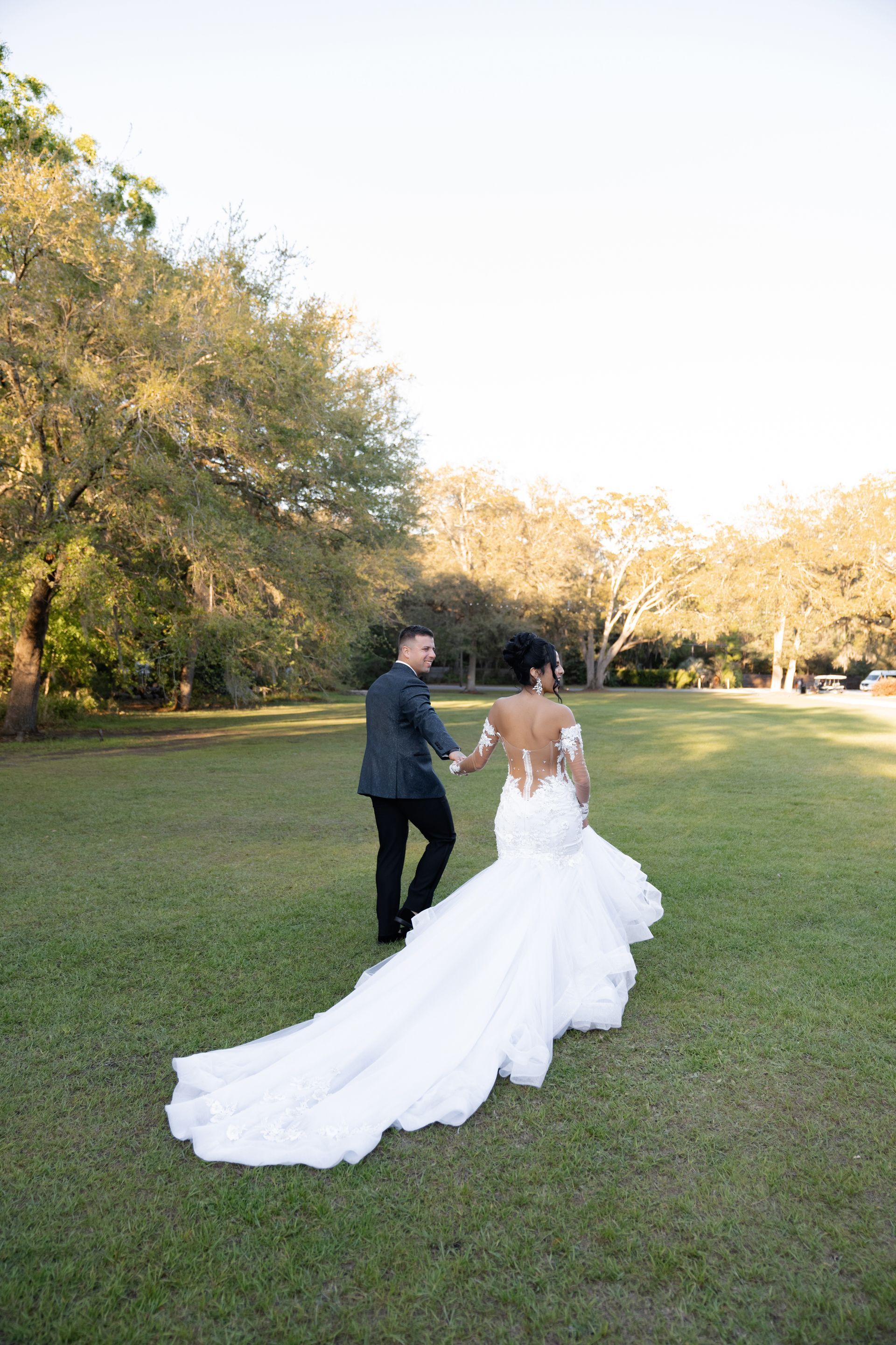 Couple in wedding attire walking across a grassy park, bride in a long white gown